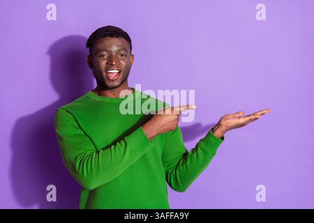 Giovane uomo in felpa verde che gestisce con le mani su sfondo viola, mostrando felicità e carisma Foto Stock