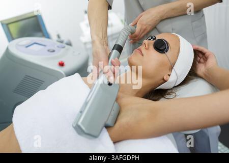Donna che riceve un trattamento laser professionale di depilazione sulle ascelle in una clinica di bellezza con occhiali protettivi. Foto Stock