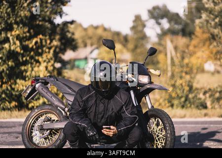 Un motociclista con l'equipaggiamento nero e il casco si siede su una motocicletta su una strada rurale in autunno Foto Stock