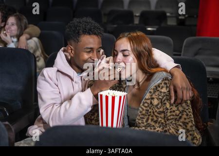 Una giovane coppia si diverte con un appuntamento al cinema, condividendo popcorn e un momento tenero nel teatro buio Foto Stock