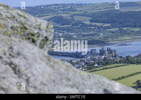 Vista dalle montagne al castello di Conwy, rovine medievali circondate da campi verdi e cielo blu, con una roccia in primo piano, Conwy, contea G. Foto Stock