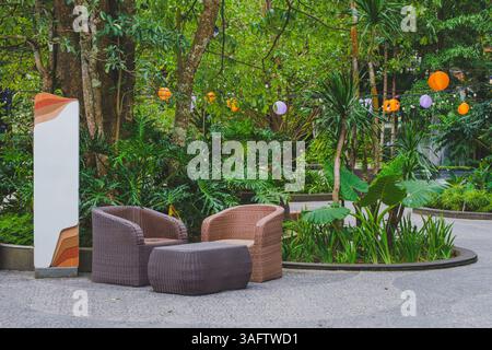 Una confortevole area lounge esterna con due sedie in rattan e un tavolo, situata in un lussureggiante giardino tropicale decorato con lanterne sospese Foto Stock