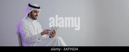 Elegante uomo saudita in abbigliamento tradizionale, che si rilassa su una comoda sedia e utilizza un tablet. L'immagine trasmette un senso di modernità e autenticità, Foto Stock