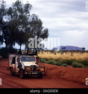 Typische Szene aus dem australischen Outback mit einer roten Sandpiste, die sich durch die trockene Landschaft zieht. Im Vordergrund fährt ein robuster Geländewagen mit zusätzlichem Equipment und Schutzbügeln durch das raue Terrain – ein typisches Fahrzeug für Expeditionen in abgelegene Gegenden. Im Hintergrund erhebt sich der markante Uluru (Ayers Rock), eine heilige Stätte der Aborigines und eines der bekanntesten Wahrzeichen Australiens. Die Landschaft ist geprägt von trockenem Grasland, vereinzelt stehenden Bäumen und einer weiten, offenen Szenerie unter bewölktem Himmel, um 1985. Foto Stock