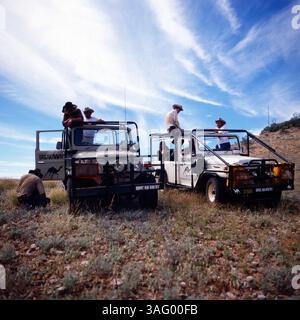 Gruppe von Abenteurern oder Outback-Reisenden, Die mit zwei robusten Geländewagen im australischen Outback unterwegs sind. Die Fahrzeuge, ausgestattet mit Schutzgittern und Expeditionsausrüstung, sind auf einer offenen Grasfläche abgestellt. Einige Personen tragen typische Outback-Hüte und Cowboy-ähnliche Kleidung, während sie sich unterhalten oder die Umgebung erkunden. Der weitläufige Himmel mit Weißen Wolken unterstreicht die Weite und Abgeschiedenheit der Landschaft, um 1985. Foto Stock