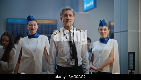 Ritratto di un pilota di linea professionale e di due bellissimi assistenti di volo in eleganti uniformi che sorridono e guardano la fotocamera. Membri dell'equipaggio di condotta nell'affollato terminal dell'aeroporto internazionale. Colpo medio. Foto Stock