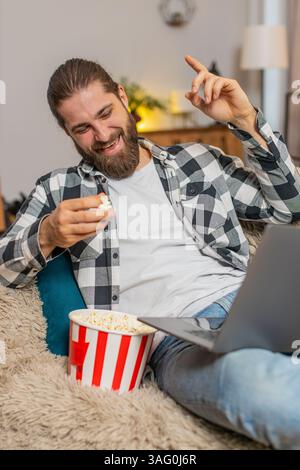 Un giovane che mangia popcorn a casa, saltando occasionalmente i chicchi giocherellando mentre guarda divertenti clip sul laptop. Un ragazzo caucasico si rilassa comodamente sul divano, ridendo ad alta voce di video sciocchi e divertenti Foto Stock