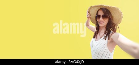 Una donna sorridente di 30-35 anni con un cappello a tesa larga e occhiali da sole su sfondo giallo, si fa un selfie. Un concetto per viaggi e blog, Foto Stock