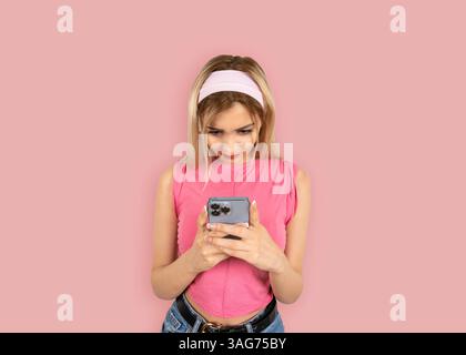 ritratto di una donna bionda sicura di sé che usa lo smartphone. Indossa una camicia rosa senza maniche che regge il telefono cellulare, digita, invia messaggi, naviga Foto Stock