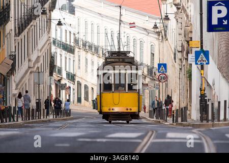 L'iconico tram giallo 28 attraversa un'affascinante strada di Lisbona, in Portogallo, circondata da edifici storici e pedoni in una tipica giornata della città. Foto Stock