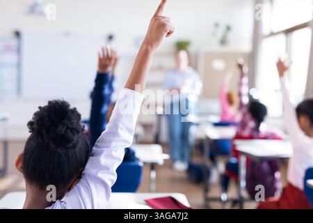 A scuola, studenti diversi che alzano la mano in classe, coinvolgendo un'insegnante femminile Foto Stock