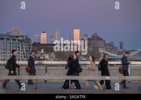 Londra, Regno Unito: Persone che camminano sul London Bridge con movimento sfocato. I grattacieli dei London Docklands riflettono il sole che tramonta. Foto Stock