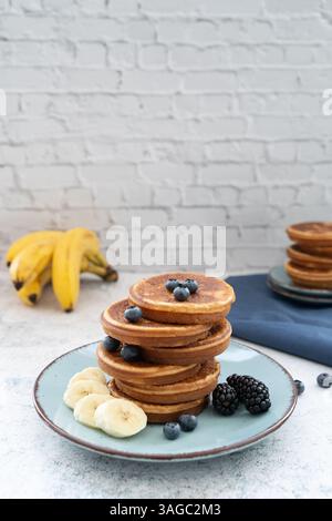 Frittelle dolci e soffici sovrapposte adornate con banana e frutti di bosco su un piatto blu. Lato con miele sul piano del tavolo grigio chiaro con un panno blu Foto Stock