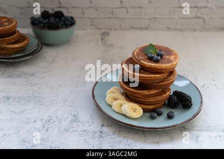 Frittelle dolci e soffici sovrapposte adornate con banana e frutti di bosco su un piatto blu. Seconda pila e ciotola di frutti di bosco sullo sfondo Foto Stock