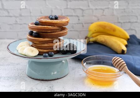 Frittelle dolci e soffici sovrapposte adornate con banana e frutti di bosco. Con il miele su piatti blu su un piano da tavolo grigio chiaro con un panno giallo Foto Stock