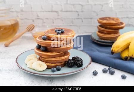 Frittelle dolci e soffici sovrapposte adornate con banana e frutti di bosco. Con il miele su piatti blu su un piano da tavolo grigio chiaro con un panno blu Foto Stock
