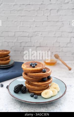 Due pile di pancake dolci e soffici adornati con banana e frutti di bosco, su piatti blu su un tavolo grigio chiaro con un panno giallo Foto Stock