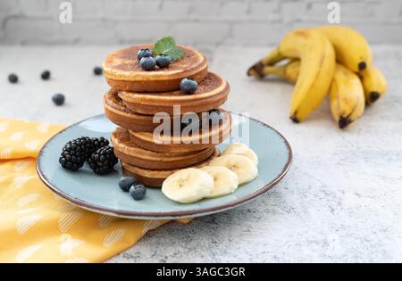 Frittelle dolci e soffici sovrapposte adornate con banana e frutti di bosco. Con il miele su piatti blu su un piano da tavolo grigio chiaro con un panno giallo Foto Stock
