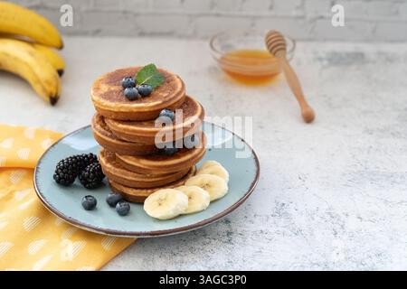 Frittelle dolci e soffici sovrapposte adornate con banana e frutti di bosco. Con il miele su piatti blu su un piano da tavolo grigio chiaro con un panno giallo Foto Stock