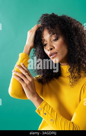 Donna con maglione giallo che tocca i capelli, esprimendo sicurezza e stile. Moda, ritratto, casual, trendy, espressione, acconciatura Foto Stock