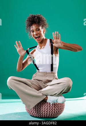 Donna allegra seduta seduta a gambe incrociate su pouf, che posa giocosamente con le bretelle. Moda, casual, donna, outfit, felicità, espressione Foto Stock