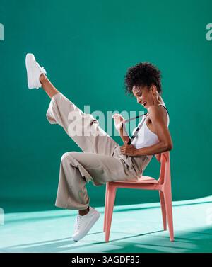 Donna allegra in un outfit casual che posa giocosamente su una sedia con uno sfondo vivace. Moda, ritratto, stile, colorato, felicità Foto Stock