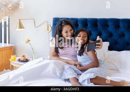Madre e figlia asiatiche sorridono mentre fanno selfie a letto a casa. Famiglia, legame, felicità, stile di vita, tecnologia, smartphone Foto Stock
