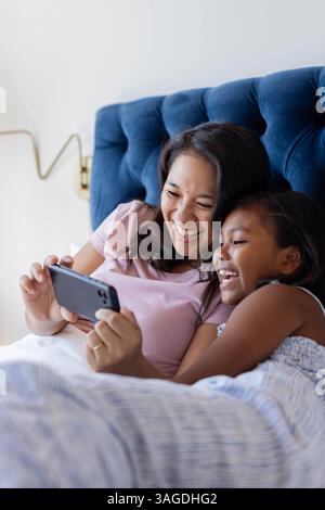 Ridendo insieme a letto, madre asiatica e figlia che guardano video sullo smartphone. Famiglia, legame, tecnologia, felicità, casa, genitorialità Foto Stock