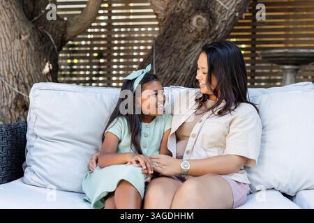 Seduti sul divano all'aperto, madre e bambino asiatici sorridono, conversano. Famiglia, legame, relax, genitorialità, connessione, tempo libero Foto Stock
