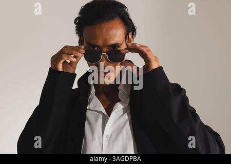 Uomo sicuro di sé che regola gli occhiali da sole indossando un elegante cappotto nero in studio di moda. stile, sicurezza, abbigliamento, uomo, alla moda, audace Foto Stock