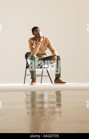 Uomo afroamericano seduto su una sedia in studio, indossa un elegante outfit casual. Moda, stile, moderno, abbigliamento casual, abbigliamento da uomo, alla moda Foto Stock
