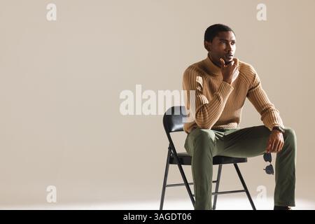 Uomo elegante in maglione seduto su una sedia, tenendo gli occhiali da sole, contemplando. Moda, stile, contemplazione, uomo, premuroso, accessori Foto Stock