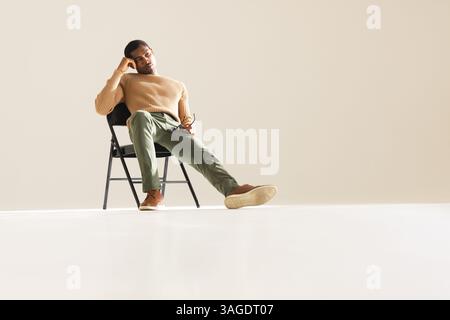 Uomo afro-americano in un elegante vestito seduto su una sedia, che sembra contemplativo. Moda, contemplazione, moderno, eleganza, posa, premuroso Foto Stock