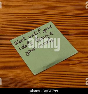 Un messaggio motivazionale scritto su una nota adesiva verde incoraggia il duro lavoro e l'ambizione. La nota è posta in modo prominente su un tavolo di legno testurizzato, sh Foto Stock