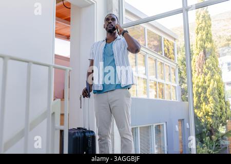 Uomo afroamericano afroamericano che tiene in mano la valigia, parla al telefono nella moderna hall dell'hotel. Viaggi, affari, comunicazione, ospitalità, bagagli, Foto Stock
