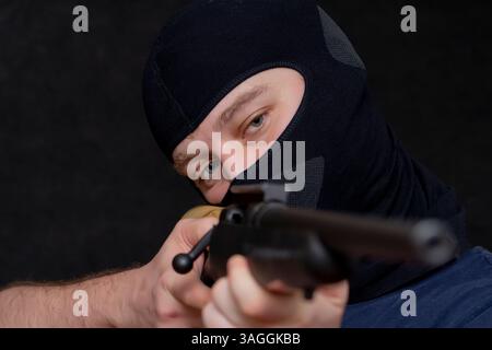 Un uomo in una balaclava punta un fucile su uno sfondo scuro e selettivo. Foto Stock