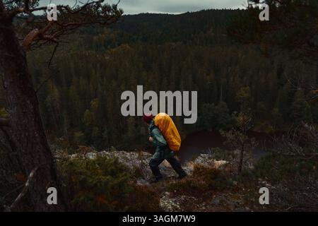 Un uomo caucasico che cammina su una scogliera che si affaccia su una foresta con uno zaino arancione. Foto Stock