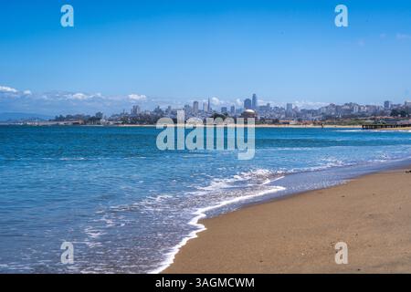 La spiaggia sabbiosa di Golden Gate Beach incontra l'Oceano Pacifico, con lo skyline di San Francisco visibile in lontananza sotto un cielo azzurro Foto Stock