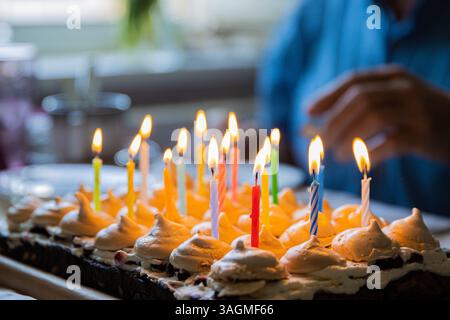 Festa di compleanno a casa. Primo piano 15 candele colorate accese sulla grande torta al cioccolato fatta in casa, decorata con meringhe e panna montata. Un uomo è S Foto Stock