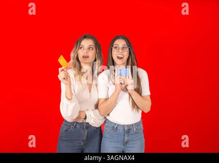 ritratto in studio rosso di due ragazze caucasiche degli anni '20 con carte di credito. Espressione del viso scioccata, due donne eccitate e allegre. Indossa una camicia bianca Foto Stock