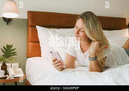 Bella donna bionda che usa lo smartphone sdraiata sul letto in camera da letto Foto Stock