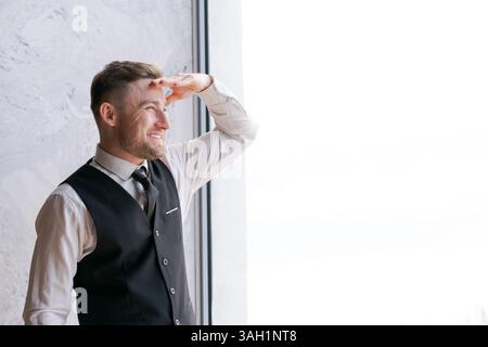 L'uomo con un gilet nero e una camicia bianca guarda fuori dalla finestra. Sta sorridendo. La finestra è chiara e la vista esterna è visibile Foto Stock