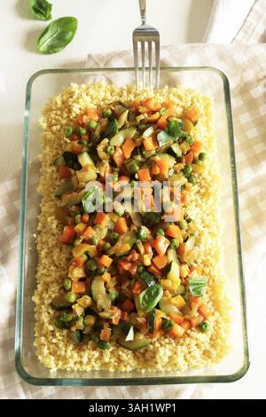 Couscous con verdure (zucchine, peperoni, carote, erbe) in un vassoio di vetro isolato su un tavolo di legno bianco. Cibo sano e vegetariano. Foto Stock