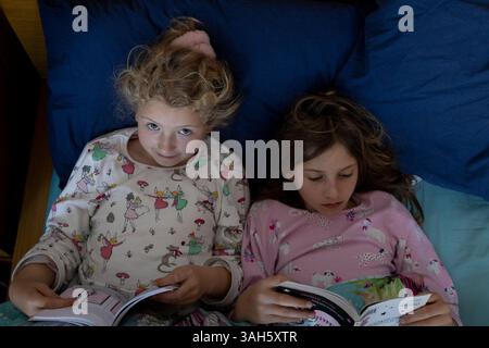 Due sorelle di bambini che leggono libri a letto fianco a fianco. Le ragazze hanno 7 anni (a sinistra) e 9. Foto Stock