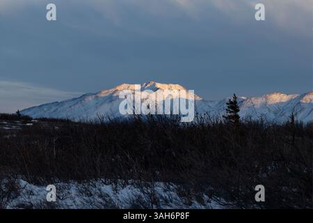 Nuvole nel cielo blu sopra la cima di una montagna innevata illuminata dal sole in una serata invernale vicino a Delta Junction, Alaska. Foto Stock