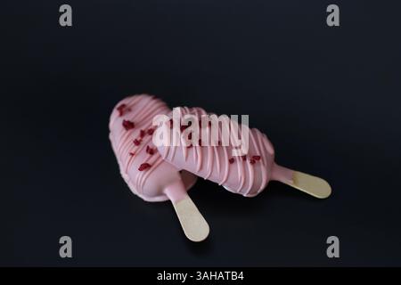 Due bacchette di gelato rosa con spruzzette rosse. I bastoncini sono posizionati su uno sfondo nero. Foto Stock