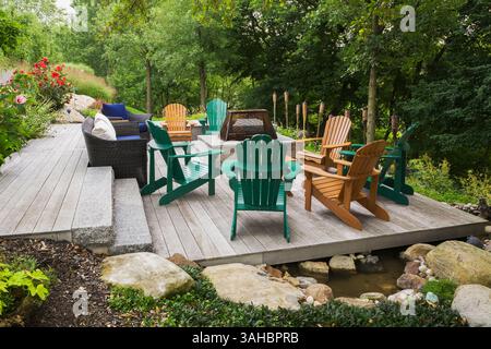 Mobili in vimini marroni, sedie Adirondack in legno verde e marrone su un terrazzo in legno grigio con focolare in un giardino in pendenza in estate. Foto Stock