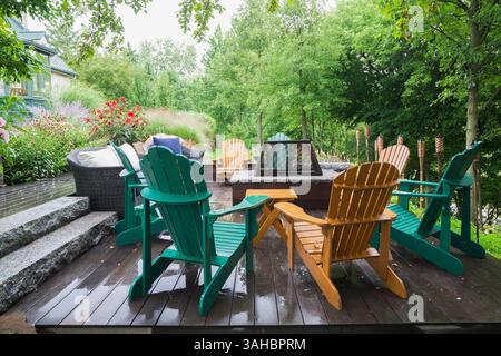 Mobili in vimini marroni, sedie Adirondack in legno verde e marrone su un terrazzo in legno grigio con focolare in un giardino in pendenza in estate. Foto Stock