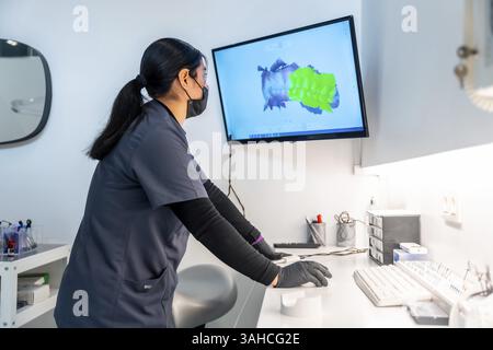 Dentista che indossa una maschera e guanti usando un computer e analizza una scansione 3d dei denti su un monitor in una clinica odontoiatrica Foto Stock