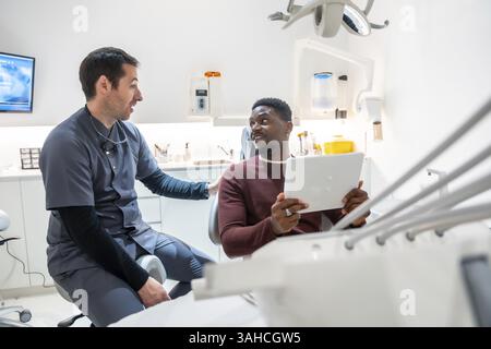 Dentista che spiega la diagnosi al paziente che tiene in mano la compressa nella clinica odontoiatrica, mostrando la tecnologia moderna e la cura del paziente Foto Stock